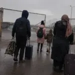A group of displaced people carrying luggage toward a closed border gate