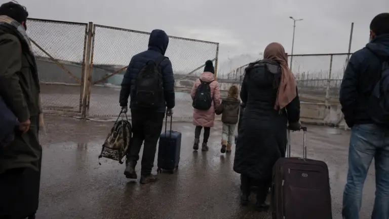 A group of displaced people carrying luggage toward a closed border gate