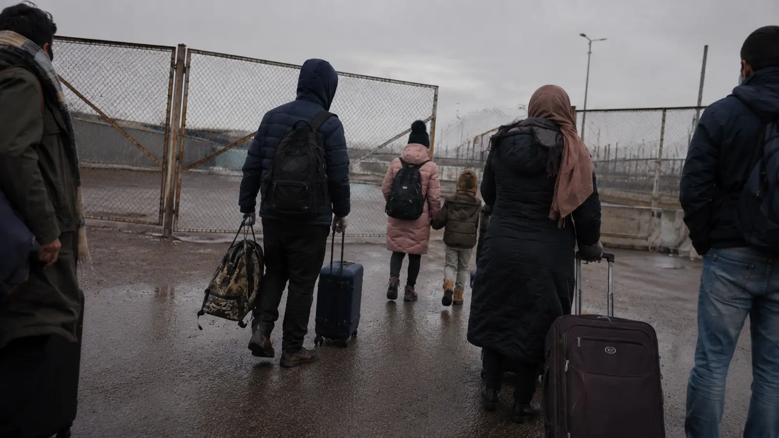 A group of displaced people carrying luggage toward a closed border gate